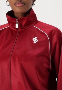 Red zip-up jacket made of smooth, lightweight material. Features white piping along the seams and a logo on the chest. High collar design.