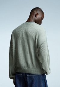 Rippstrickpullover in Hellgrün mit Rundhalsausschnitt und überschnittenen Schultern, kombiniert mit dunkelblauen Jeans, sichtbare Texturdetails.