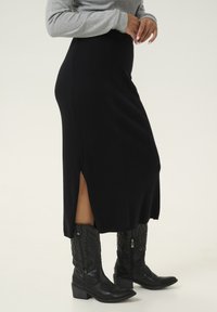 Fustă midi neagră din tricot cu nervuri și fanta laterală, asociată cu cizme cowboy negre cu imprimeu. Partea de sus este un pulover ajustat de culoare gri deschis.