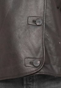 Nærbillede af en mørkebrun læderjakke med to sorte knapper og synlig syning over mørk denimstof.