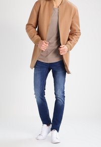 Beige bomberjacka med ribbade muddar, bärs över en ljusgrå skjorta. Blå jeans och vita sneakers fullbordar outfiten. Neutral bakgrund.