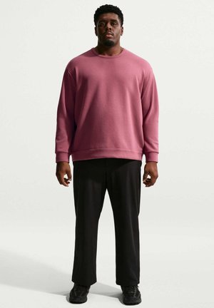 Un uomo adulto è in piedi di fronte a noi, indossa una felpa rosa a maniche lunghe, pantaloni neri e scarpe da ginnastica nere su uno sfondo bianco semplice.