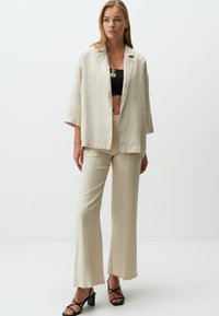 Beige Leinenblazer über einem schwarzen Croptop, kombiniert mit beigen Schlaghosen. Schwarze Blockabsatzsandalen runden das Outfit ab. Eine schlichte goldene Halskette ist ebenfalls enthalten.