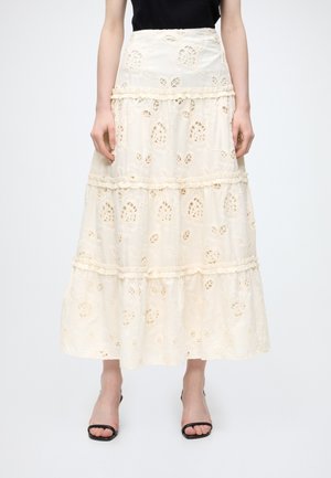 Jupe midi en dentelle blanche avec des découpes florales et des volants à étages, portée avec des sandales noires à brides et un haut noir.