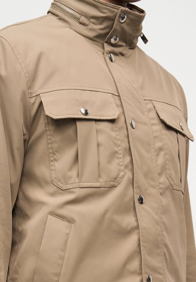 Veste beige à col montant, fermeture éclair et boutons-pression, deux poches poitrine à rabat avec boutons, et poche latérale cousue.