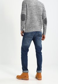 Pull texturé gris avec des patchs coudes foncés, jean bleu et bottes marron. Le jean est retroussé aux chevilles, dévoilant les bottes.