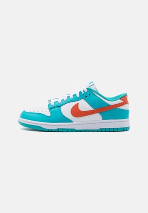 Low-top sneaker met witte basis, turquoise overlays, oranje Nike-swoosh, witte veters en turquoise buitenzool op een effen achtergrond.