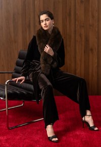 Femme portant une tenue en velours sombre et une écharpe en fourrure assise sur une chaise en cuir noir devant un mur en panneaux de bois et un tapis rouge.