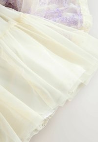 Tulle beige chiaro stratificato con finitura texturizzata, caratterizzato da dettagli floreali lilla e bordatura in pizzo lungo i bordi.