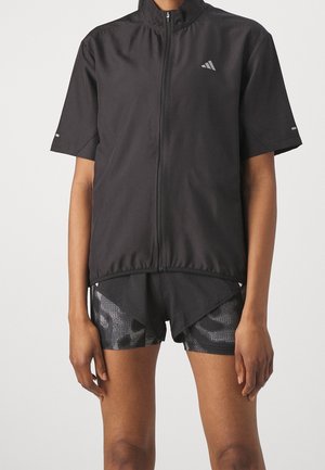Personne portant une veste noire à manches courtes zippée par-dessus un short de sport noir à motifs, debout devant un fond clair uni.