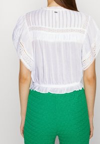 Blusa bianca leggera con maniche corte, dettagli in pizzo e vita arricciata. Abbinata a pantaloni verde a vita alta con texture.