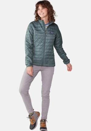 Chaqueta acolchada verde con cremallera frontal, mangas largas y cuello alto, combinada con pantalones grises y botas de senderismo marrones.