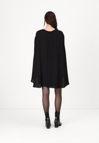 Črna obleka s plaščem in tekočo silhueto, okroglo izrezom in detajlom s ključavnico na hrbtu. Ustrezno kombinirana s prosojnimi črnimi nogavicami in gležnjarji.