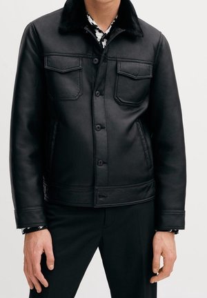 Homme portant une veste en cuir noire boutonnée avec poches poitrine à rabat et un pantalon noir devant un fond uni.