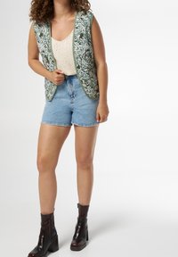 Gilet verde ricamato sopra una maglietta beige, abbinato a pantaloni shorts di jeans. Stivaletti con tacco marroni. Il gilet presenta motivi floreali e tasche.