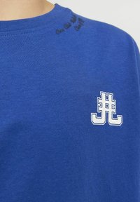 Blauwe katoenen T-shirt met een wit logo op de linkerborst en zwarte tekst bovenaan de ronde hals. Soepele textuur met een klassieke pasvorm.