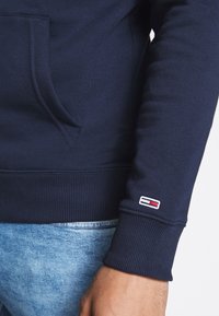 Granatowa bluza z długim rękawem z małym haftowanym logo w czerwono-biało-niebieskich kolorach w pobliżu nadgarstka, w zestawieniu z jasnoniebieskimi dżinsami.