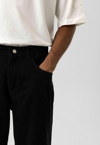 Zwarte denim broek met een ontspannen pasvorm, voorzien van zijzakken en een zichtbare metalen knoop. Gecombineerd met een loszittend wit katoenen T-shirt.