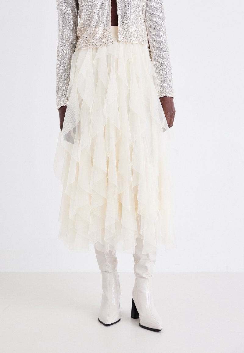 Mannequin à la peau foncée portant une jupe en tulle crème en plusieurs couches, une veste argentée à sequins et des bottes blanches brillantes montantes jusqu’aux genoux avec talons carrés.