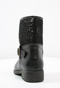 Botín negro con una base de cuero texturizado y una parte superior de tela de encaje con patrones circulares. Incluye un hebilla decorativa en el lateral.