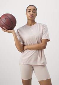Šviesiai rožinė medvilninė marškinėliai su Jordan logotipu, derinami su šviesiai rožiniais dviratininkų šortais. Modelis laiko rudą krepšinį su logotipo detale.