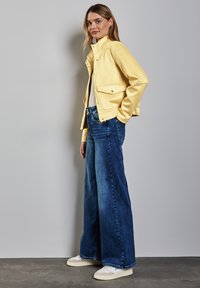 Veste en cuir jaune avec poches latérales, portée sur un haut blanc. Un jean en denim bleu à jambe large et des baskets beiges complètent la tenue.