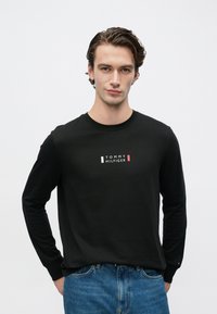 Black long-sleeve t-shirt made of cotton featuring a white and red "TOMMY HILFIGER" logo centered on the chest. Worn with blue denim jeans.