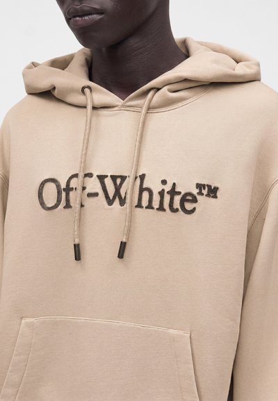 Personne portant un sweat à capuche beige avec cordons et texte brodé "Off-White™" sur la poitrine, montrant le haut du torse et le cou.