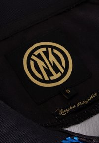 Zwarte stoffen label met een ronde gouden logo, met onderling verbonden letters. Bevat een etiket met "Gerecycled Polyester" en maat "S."