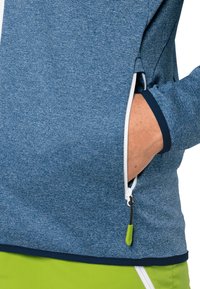 Hand i fickan på en blå zipparjacka med grön dragkedja och limegröna byxor synliga nedanför.