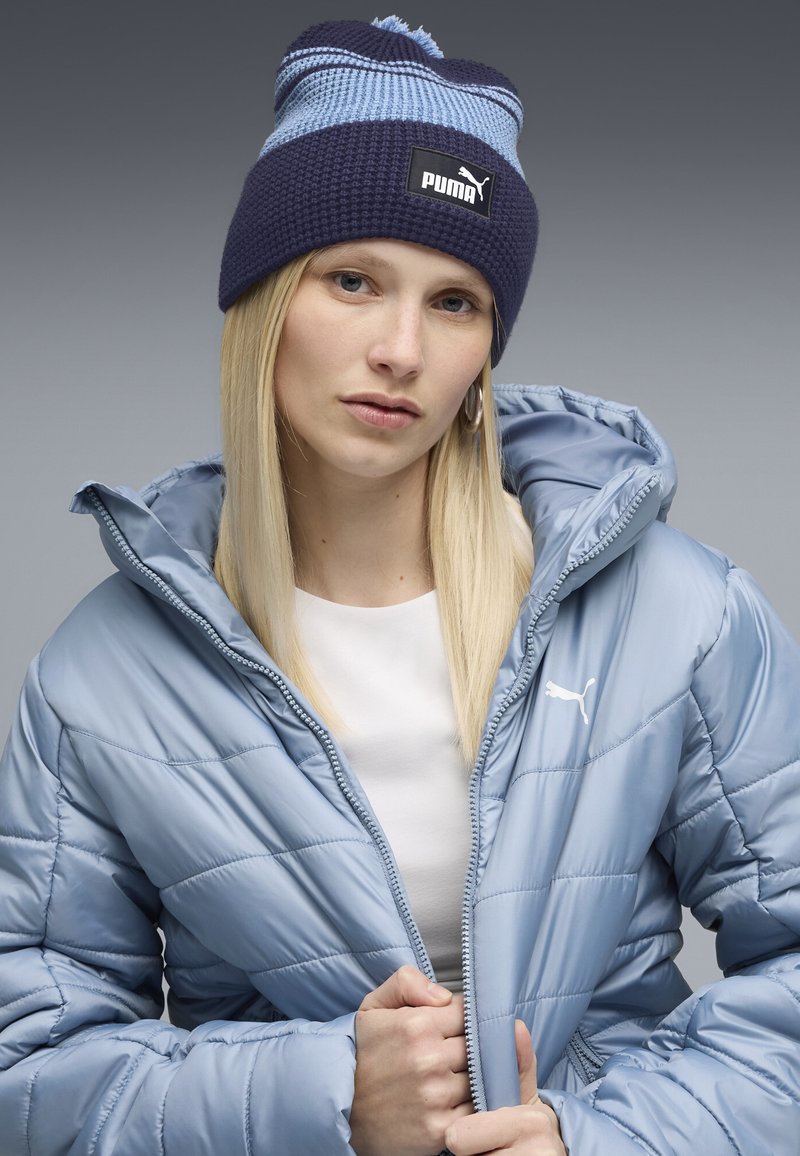 Blauwe gewatteerde jas met rits, met een logo. Marineblauwe en lichtblauwe gestreepte gebreide muts met een pompon en logopatch.