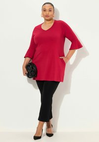 Top tunica rossa con scollatura a V, maniche a campana 3/4 e tasche laterali, abbinato a pantaloni neri slim-fit e tacchi neri. Texture liscia.