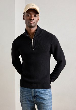 Jersey negro de punto con cierre, textura acanalada, cuello alto y detalle de cremallera plateada, combinado con jeans de mezclilla azul claro. Una gorra beige añade contraste.