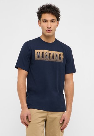 Junger Mann mit lockigem Haar, der ein marineblaues T-Shirt mit dem "MUSTANG"-Logo und beige Hosen vor einem einfarbigen hellen Hintergrund trägt.