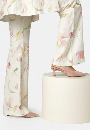 Beine in cremefarbenen, geblümten, weiten Hosen und rosa Wildleder-Slingback-Schuhe mit kleinem Absatz, wobei ein Fuß auf einem runden Sockel ruht.