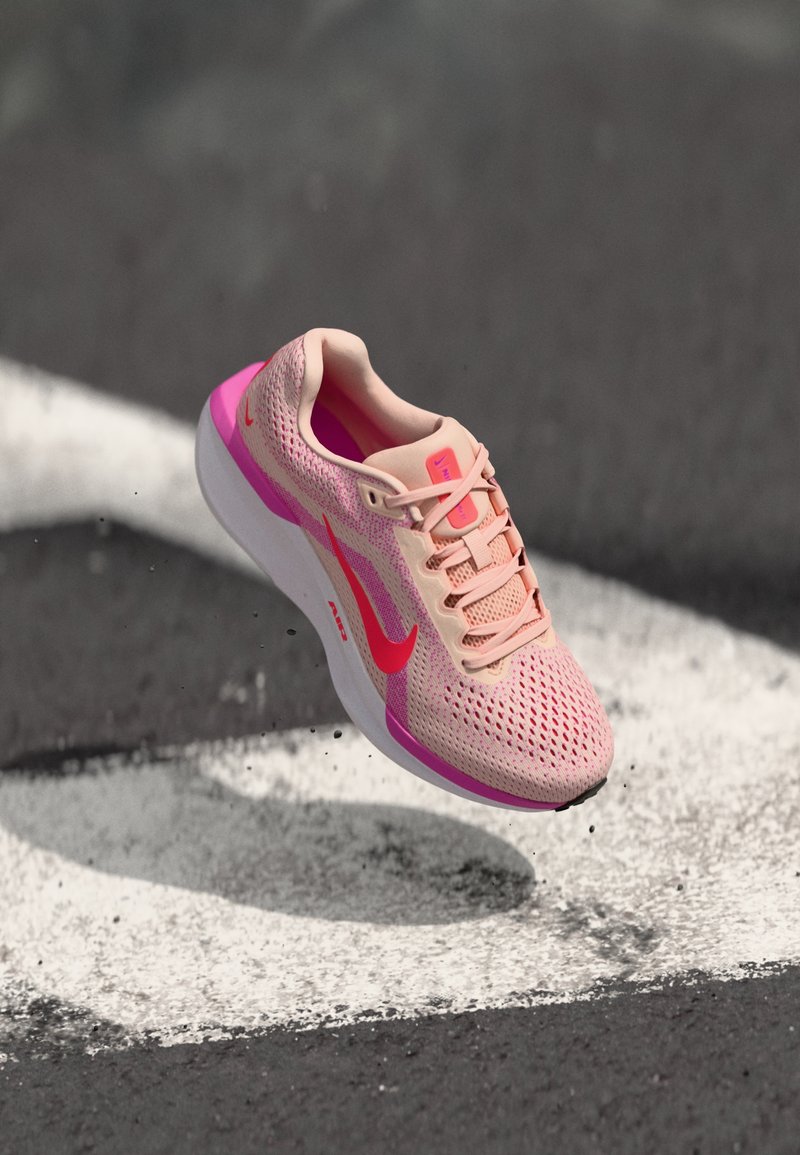 Lichtroze sport schoen met mesh bovenwerk, roze accenten en witte zool. Heeft een ronde vorm en platte veters. Nike-logo in felrood.