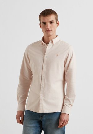 Giovane uomo con indosso una camicia beige chiara con bottoni e maniche lunghe e jeans blu, in piedi davanti a uno sfondo bianco neutro.