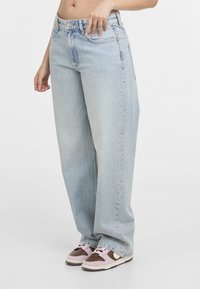 Personne portant un jean large bleu clair avec des détails subtils en strass sur les côtés et des baskets marron et roses, vue de la taille jusqu'aux pieds.