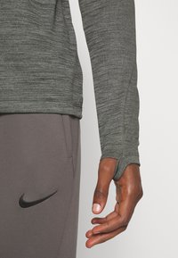 Haut de sport à manches longues gris en tissu texturé, présentant des ouvertures pour les pouces et une coupe ajustée, associé à un pantalon gris foncé avec un logo noir.