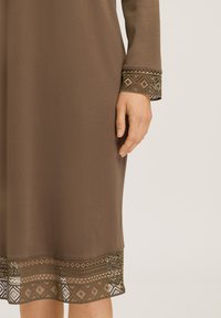 Brun klänning med långa ärmar, en texturerad nederdel med geometriska utskärningar och mönstrade detaljer i mörka och ljusa nyanser.