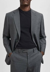 Veste de costume grise sur mesure associée à un t-shirt noir à col rond. Le costume présente un revers cranté et un pantalon à plis plats.