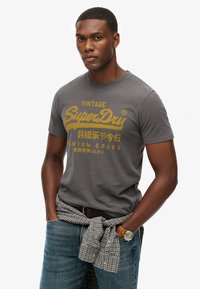 Graues Baumwoll-T-Shirt mit gelbem "Vintage SuperDry"-Logo, Rundhalsausschnitt, kurze Ärmel, kombiniert mit blauen Jeans und einem karierter Hemd, das um die Taille gebunden ist.