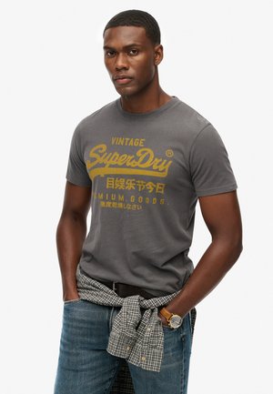 Gray cotton t-shirt with yellow "Vintage SuperDry" logo, round crew neck, short sleeves, paired with blue jeans and a checkered shirt tied at the waist.