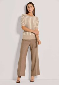 Vrouw in een beige, verticaal gestreepte, korte mouw trui, met een brede taupe broek en zwarte open teen hakken, staande tegen een effen achtergrond.