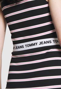 Černý a růžový pruhovaný tílko s širokým elastickým pasem, na kterém je v černé barvě vytištěno "TOMMY JEANS". Měkká, přiléhavá textilie.
