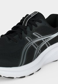 Μαύρο παπούτσι για τρέξιμο Asics με λευκές και γκρίζες λωρίδες λογότυπου, μαύρα κορδόνια και παχιά λευκή σόλα σε ανοιχτό φόντο.