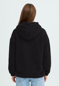 Hoodie en polaire noir avec une texture douce, coupe surdimensionnée et grande capuche. Équipé de poignet et d'ourlet élastiqués. Coutures arrière visibles.