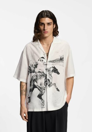 Joven con cabello largo que lleva una camisa blanca de manga corta y holgada con un boceto negro de un caballo y jinete, combinada con pantalones negros.