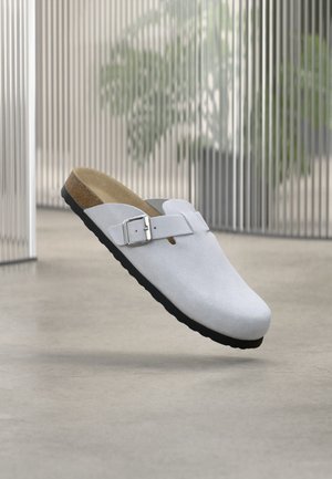 Sabot blanc à enfiler avec boucle argentée réglable, semelle intérieure en liège et semelle noire, flottant à l'intérieur sur un sol lisse près de stores verticaux.