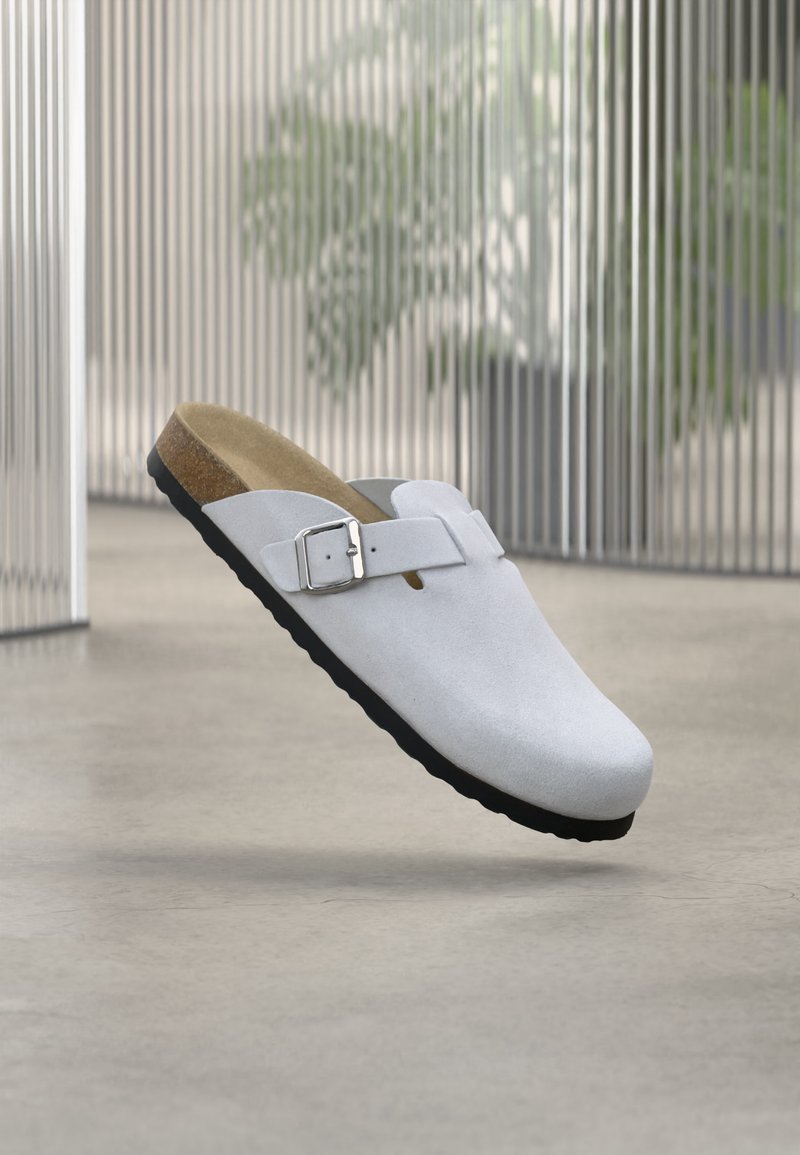 Λευκός κλειστός πάτος με ρυθμιζόμενο ασημένιο πόρπη, πάτο από φελλό και μαύρη σόλα που αιωρείται σε εσωτερικό χώρο πάνω σε λείο πάτωμα κοντά σε κάθετες περσίδες.